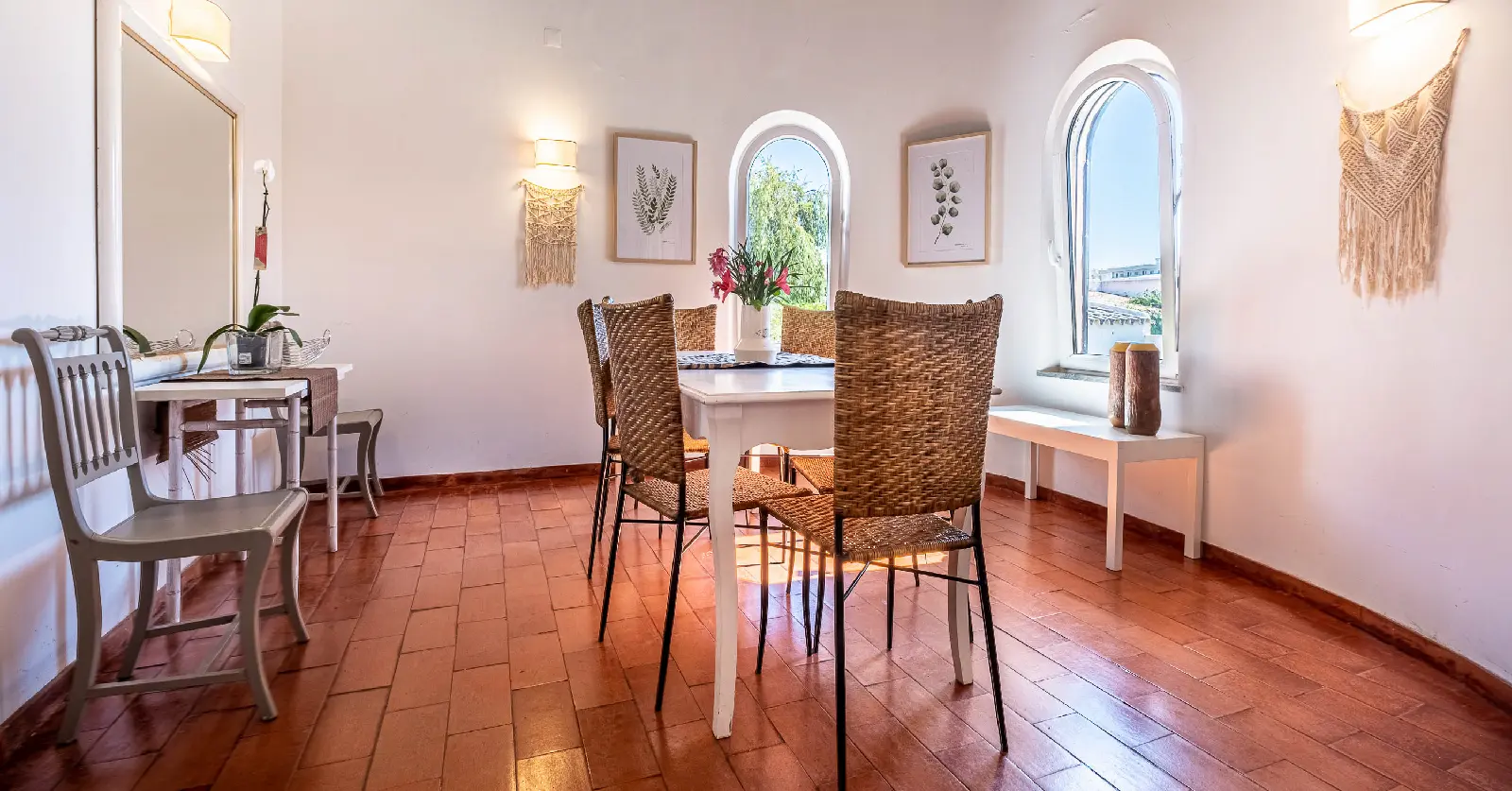 Sala de refeições com decoração boho e luz natural, com cadeiras em verga, mesa branca, janelas arqueadas e elementos decorativos em macramé — ideal para representar transformação de espaços interiores com charme rústico.
