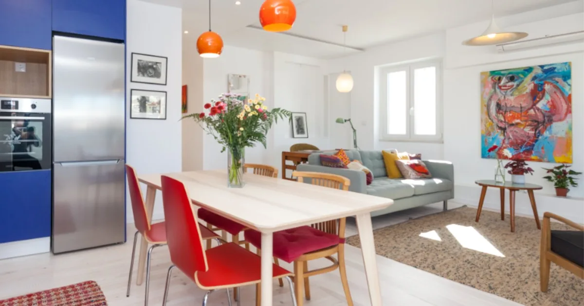 Sala de estar colorida com cozinha integrada, mesa de jantar clara, pendentes laranja e arte contemporânea na parede